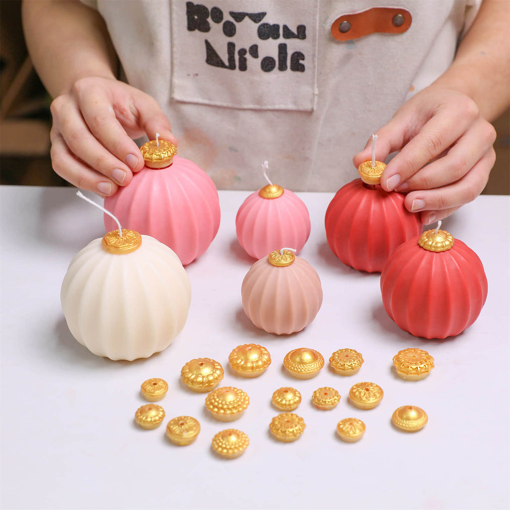 Hands assembling fluted bauble candles in ivory, blush, and red with interchangeable gold topper caps—multiple sizes on worktable - Boowan Nicole