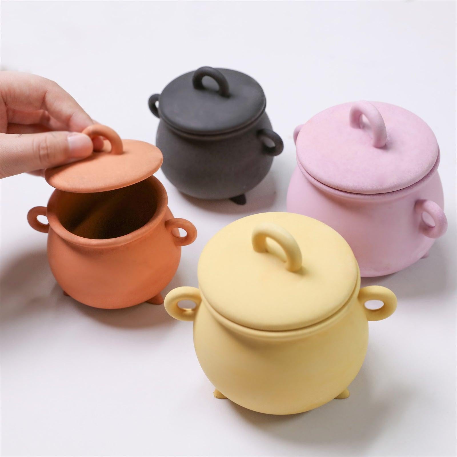 Four colorful cauldron-shaped candle jars with lids in orange, yellow, pink, and black, one being opened by hand - Boowan Nicole