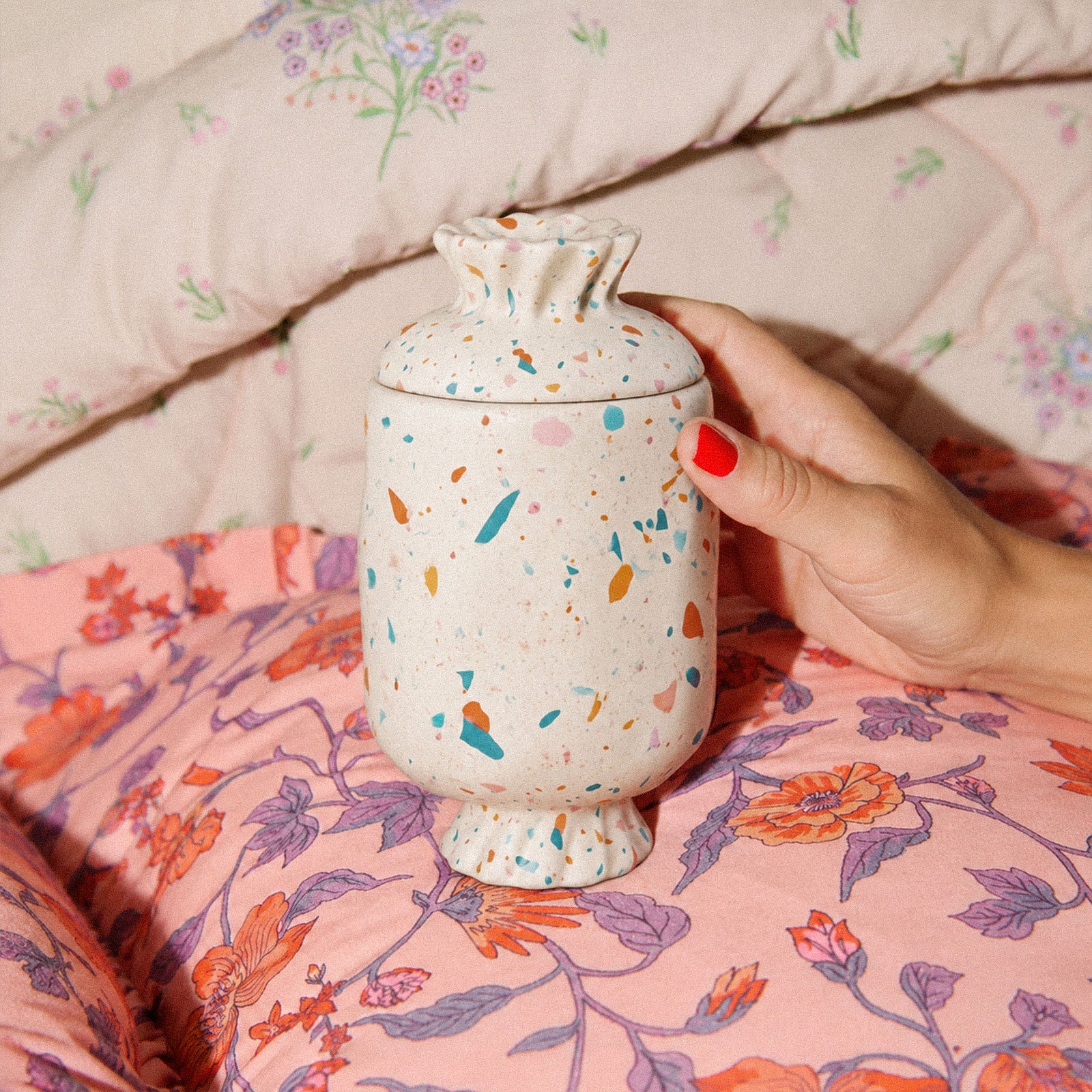 Hand holding terrazzo wrapped bonbon lidded candle jar on floral printed bedding cozy bedroom lifestyle shot - Boowan Nicole