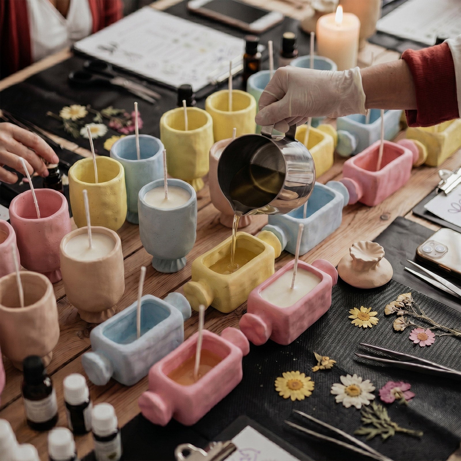 DIY candle workshop scene: pouring melted wax into assorted pastel wrapped bonbon candle blanks wooden craft table - Boowan Nicole
