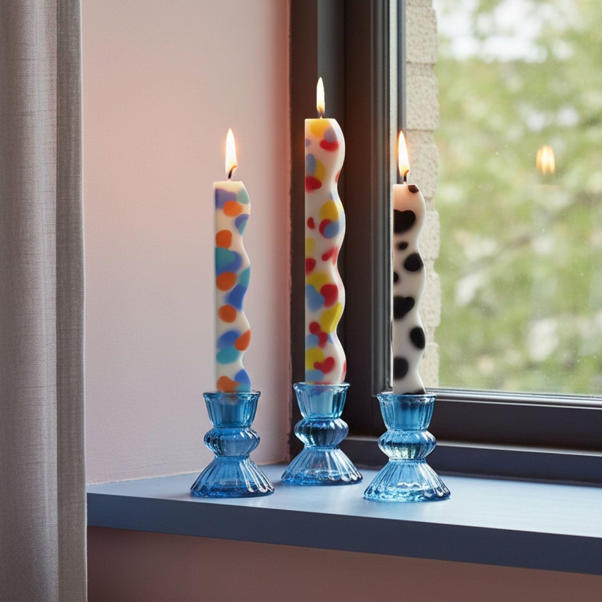 Three zigzag taper candles with colorful spots in blue glass holders on a windowsill - Boowan Nicole