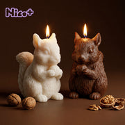 Two Acorn Hug Squirrel Candles burning side by side, one ivory and one brown, each holding a walnut, with whole and cracked walnuts scattered on a dark brown surface, Nico+ collaboration logo in the corner.