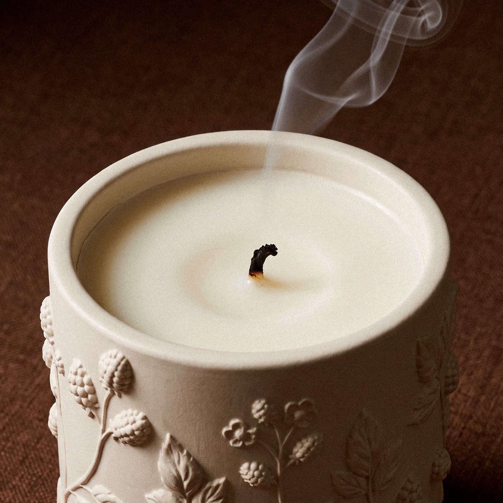 Close-up top-down view of a cream Berry Vine Candle Jar showing the wax pool, an extinguished wick with rising smoke, and the berry vine relief texture around the rim.