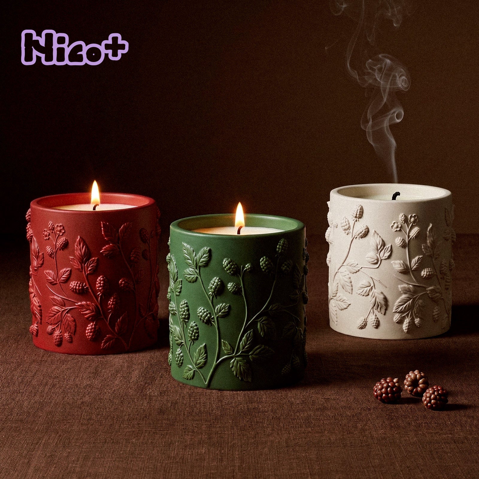 Three Berry Vine Candle Jars burning side by side in red, green, and cream, each wrapped in a berry vine relief, with blackberries scattered on a brown fabric surface and the Nico+ collaboration logo in the corner.