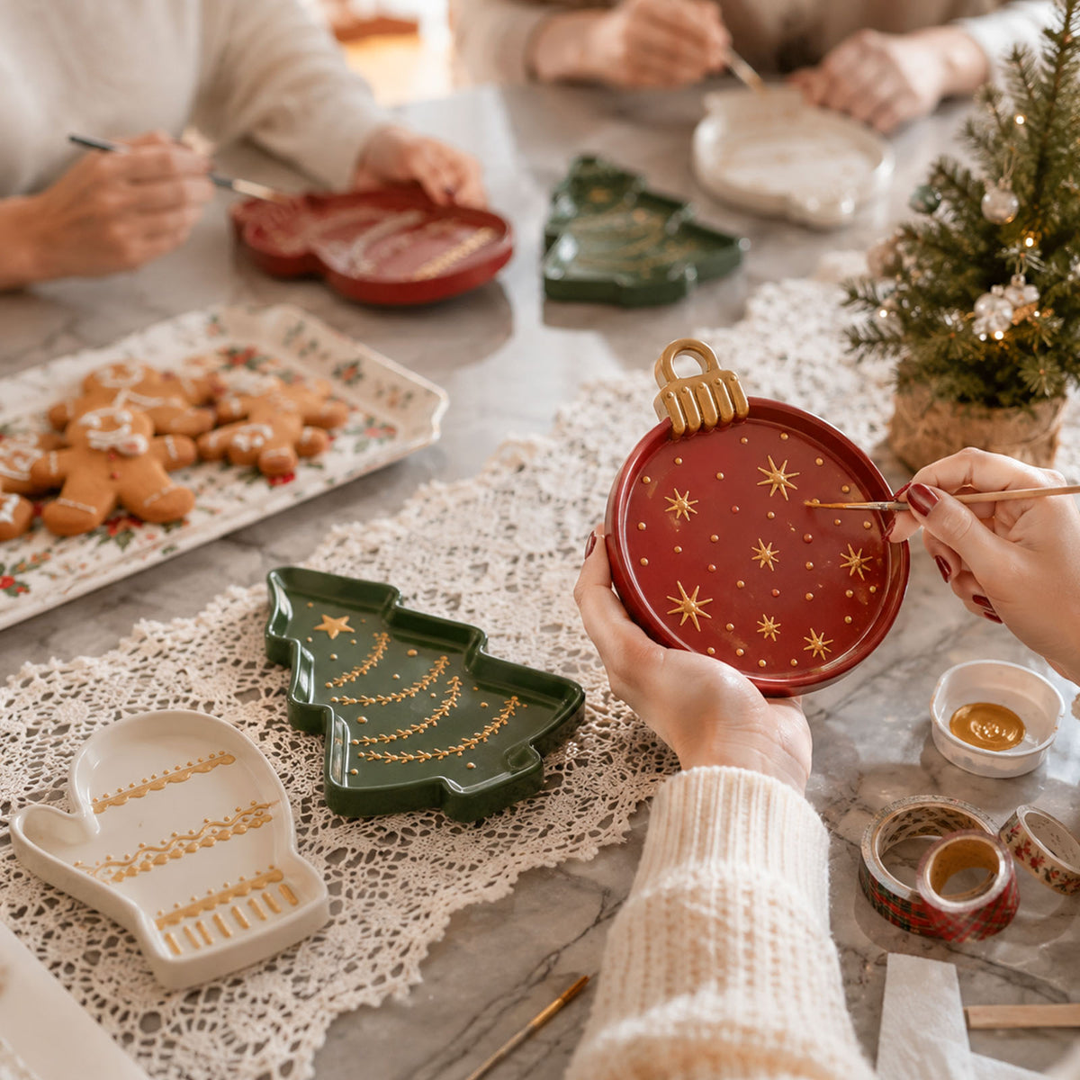 Friends painting gold details on Christmas trays at a festive table with gingerbread cookies