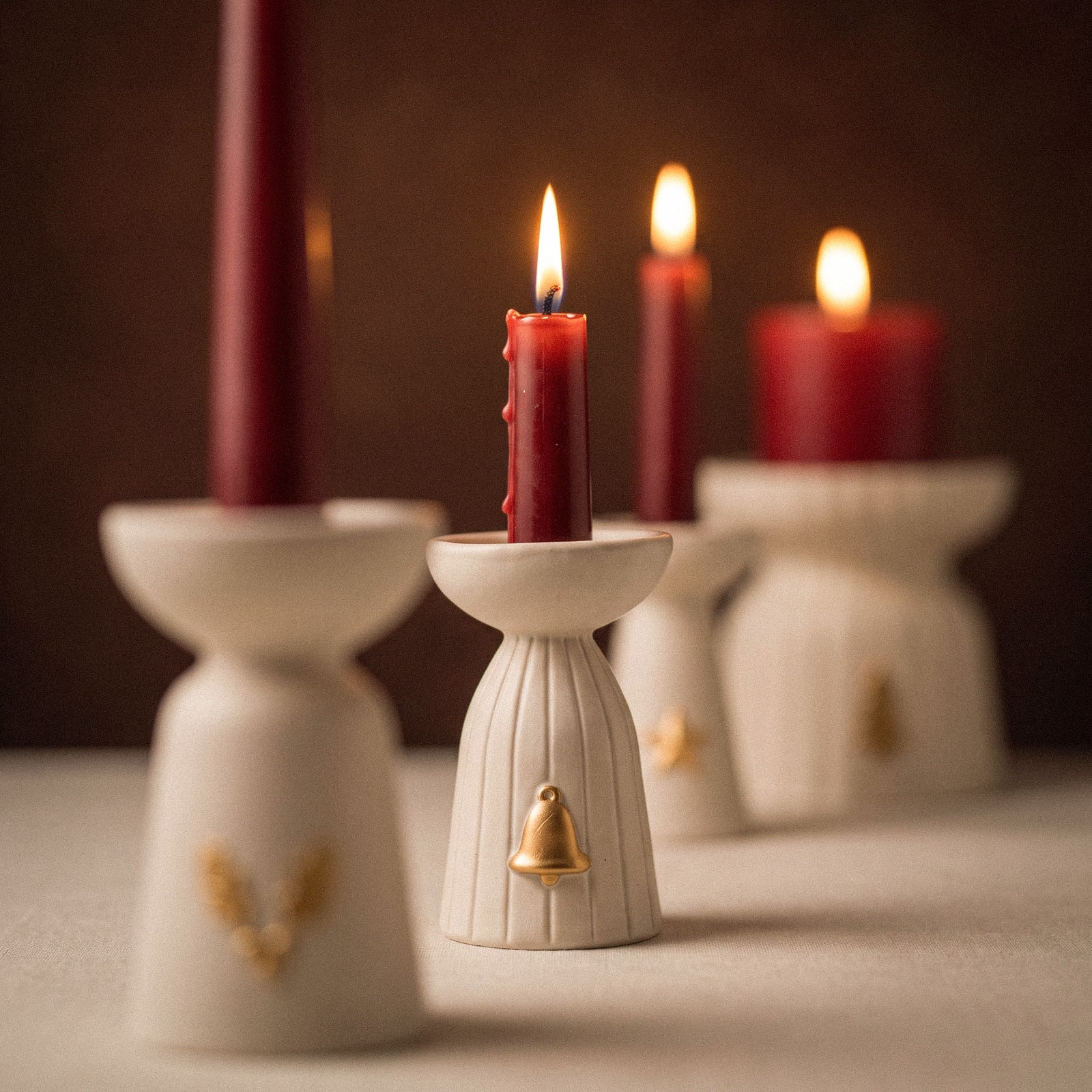 Close-up of a lit red taper candle in the bell charm Christmas Glow candle holder, with the holly, star, and tree charm holders softly blurred behind.