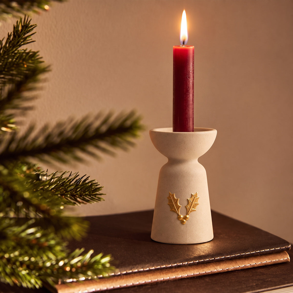 Holly charm taper candle holder with a lit red candle on a leather book beside a Christmas tree