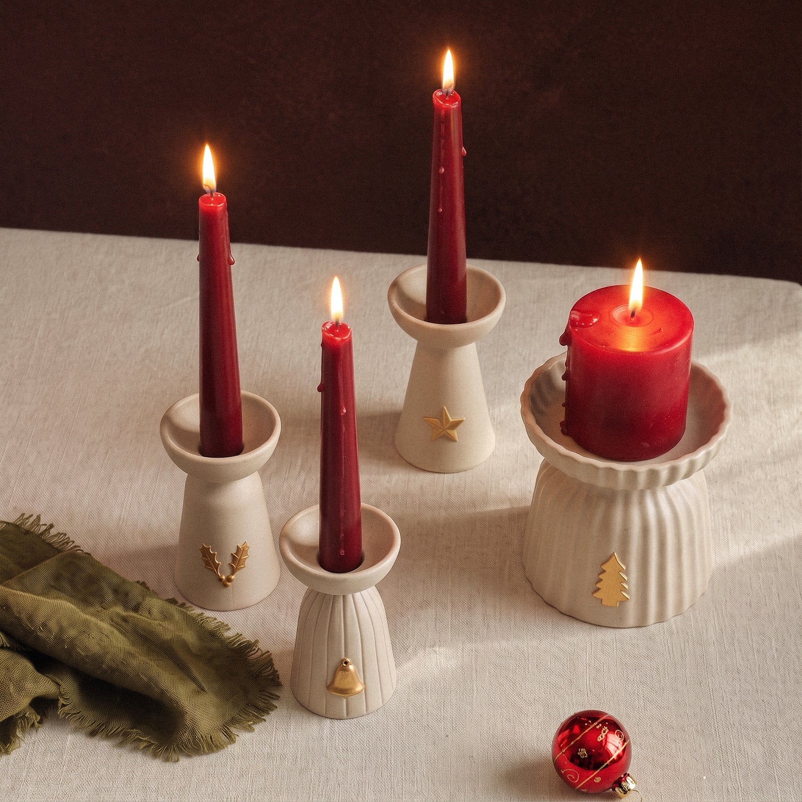 Four lit red taper and pillar candles in Christmas Glow ceramic-look candle holders on a linen tablecloth with a green napkin and a red glass ornament.