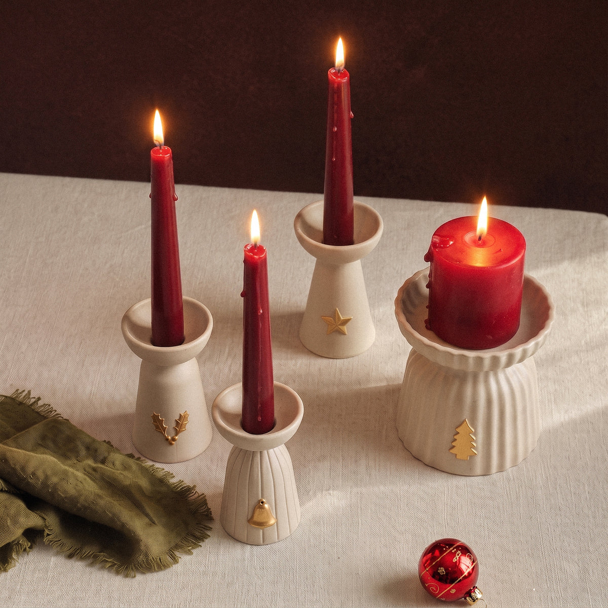 Four lit red taper and pillar candles in Christmas Glow ceramic-look candle holders on a linen tablecloth with a green napkin and a red glass ornament.
