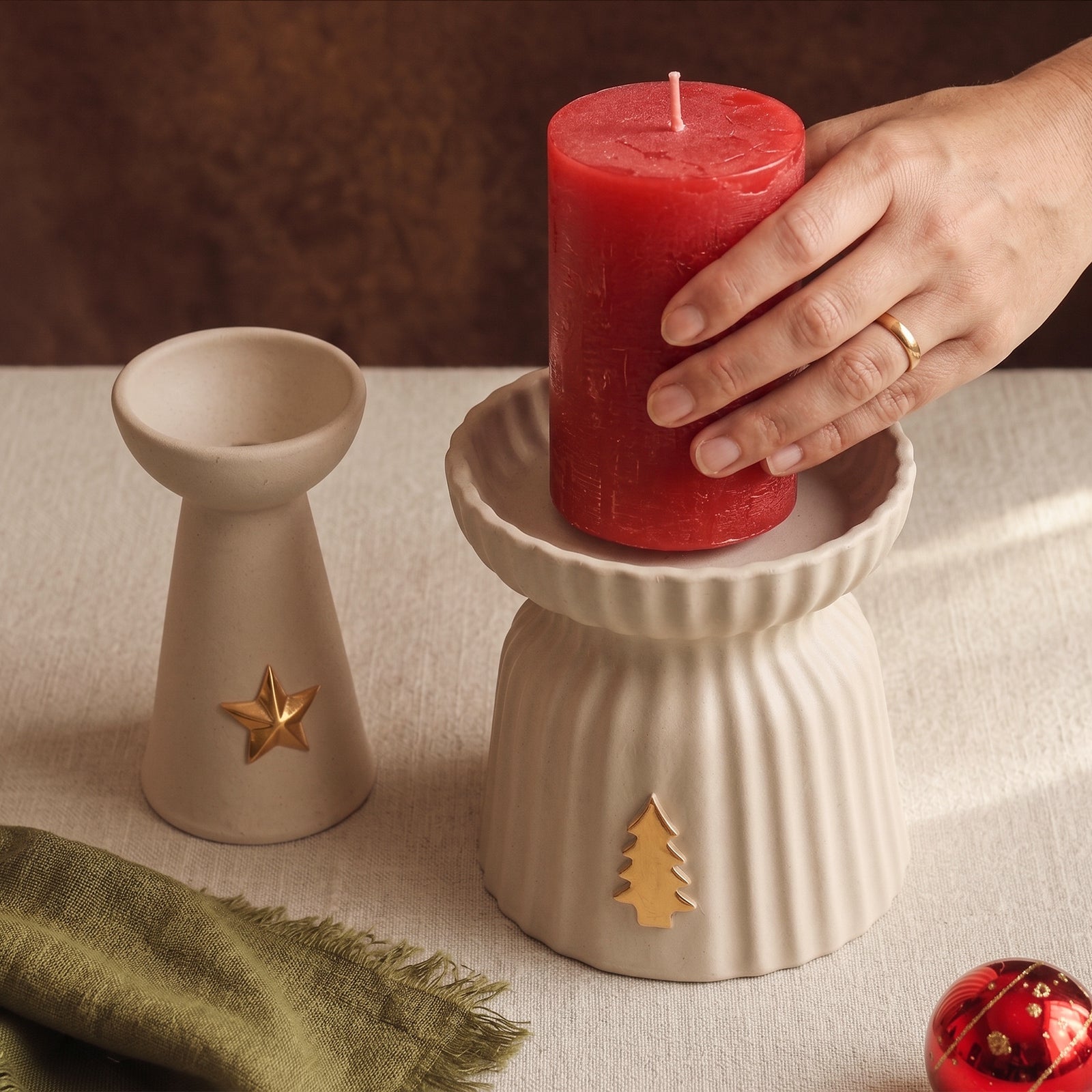 A hand setting a red pillar candle into the large ribbed Christmas Glow candle holder with a gold tree charm, beside the star charm taper holder.