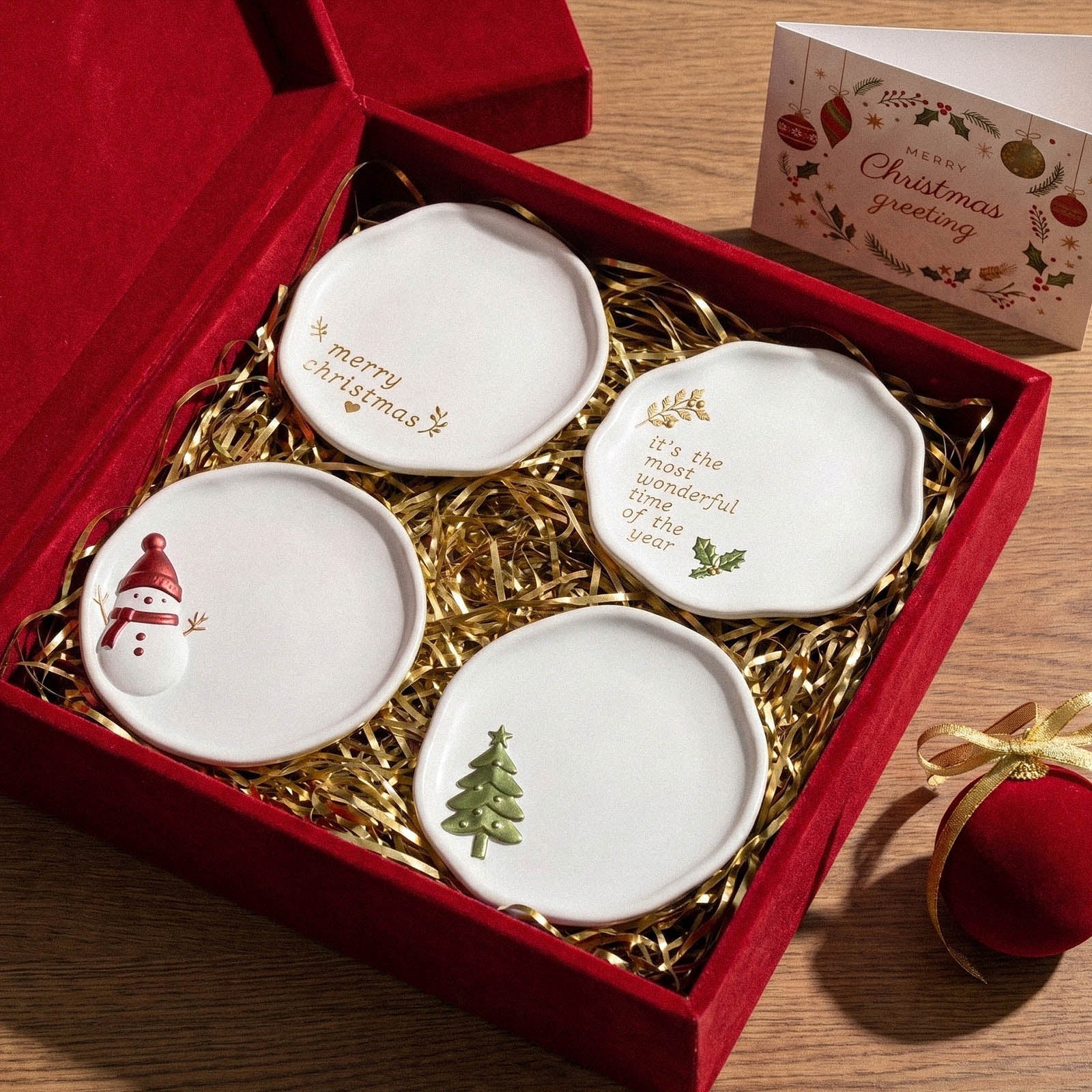 Four Christmas trinket trays nested in a red gift box lined with gold shred, next to a Merry Christmas greeting card and a red velvet ornament.