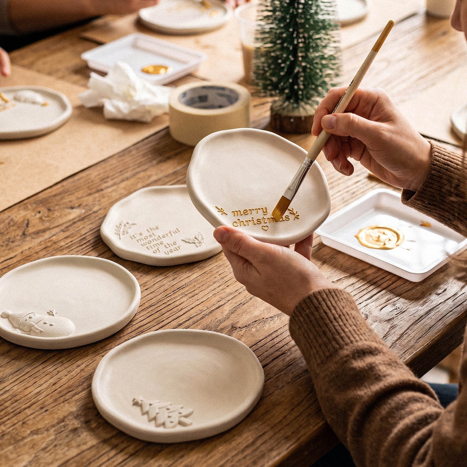 An artisan hand-painting gold Merry Christmas lettering onto a white ceramic-look trinket tray with a fine brush, with other Christmas trays arranged on a wood table.