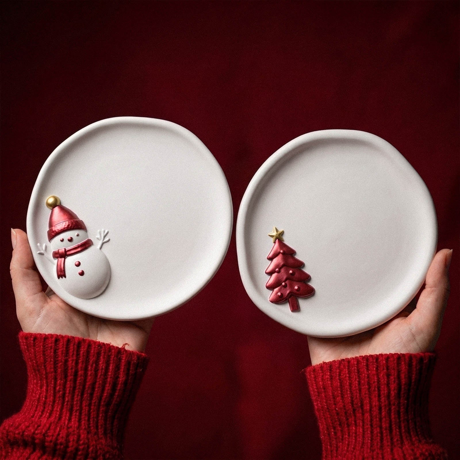 Two hands holding up a snowman-embossed trinket tray and a Christmas tree-embossed trinket tray against a red velvet backdrop, styled with a red knit sweater.