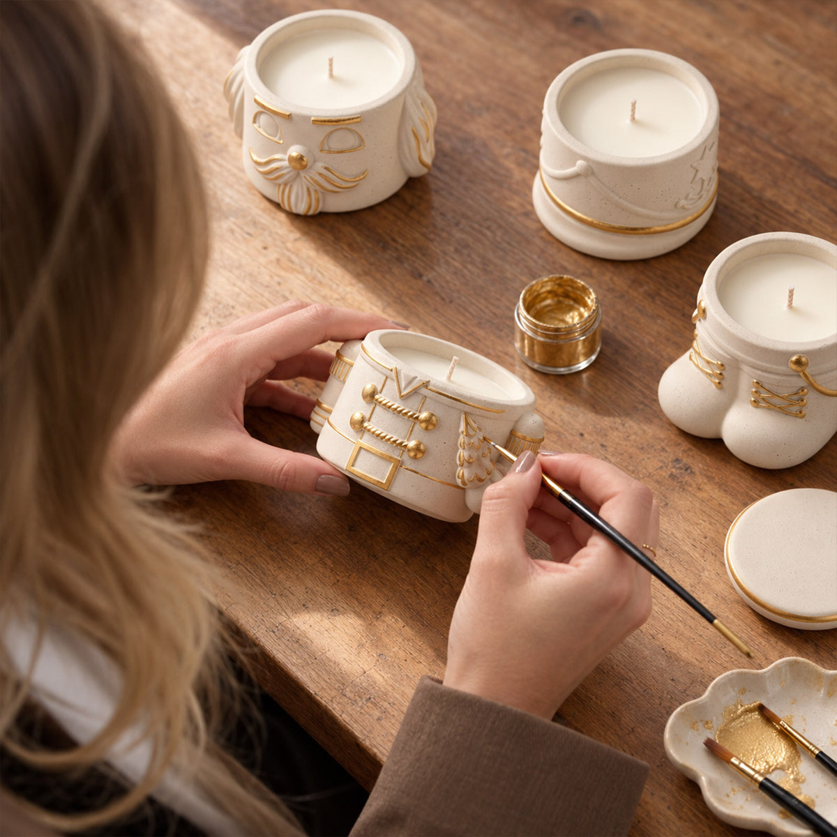 Painting gold details on cream nutcracker candle jars with fine brushes