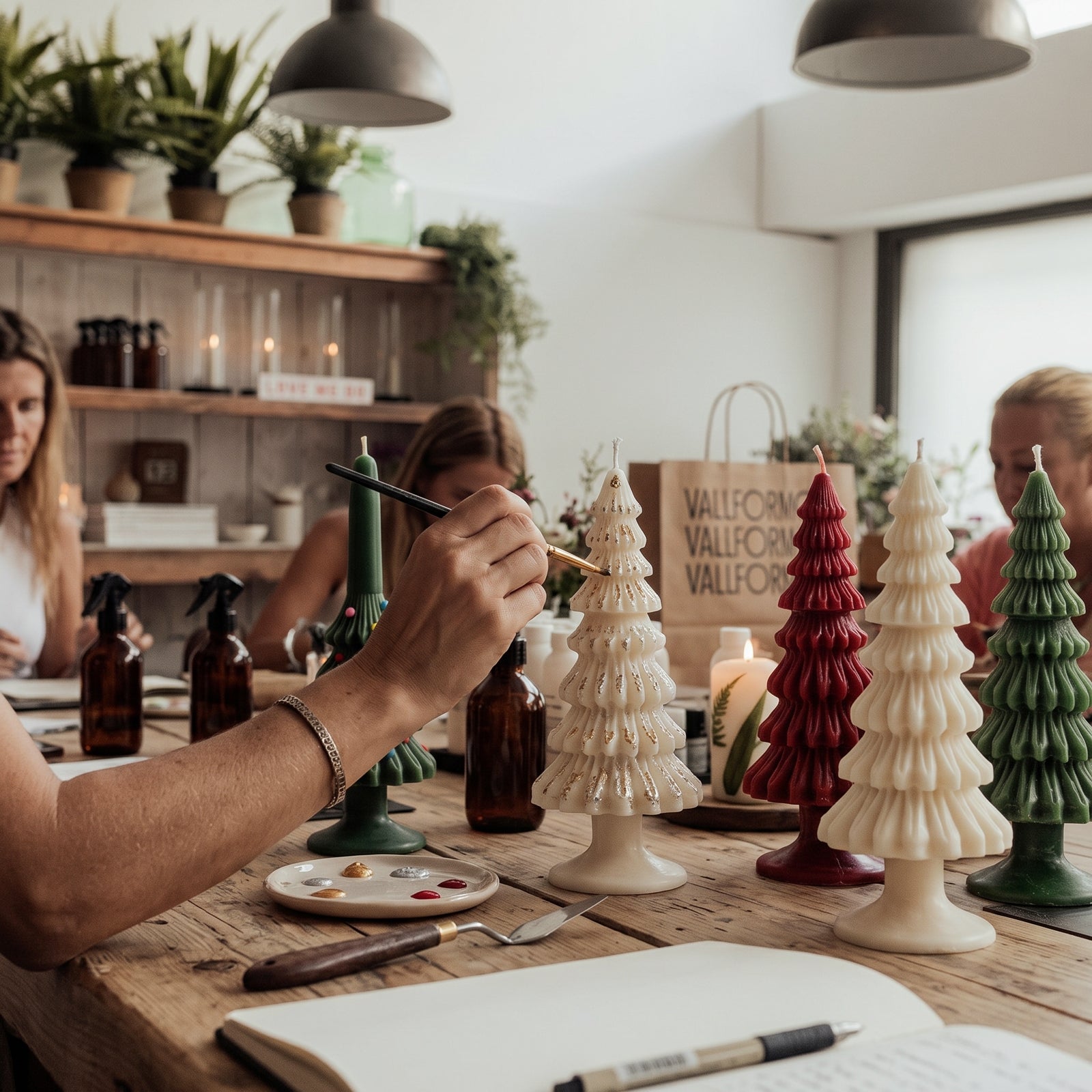 A crafter hand-painting gold accents onto a cream Christmas Tree Taper Candle in a workshop, with red, cream, and green tree candles lined up on a wooden table.
