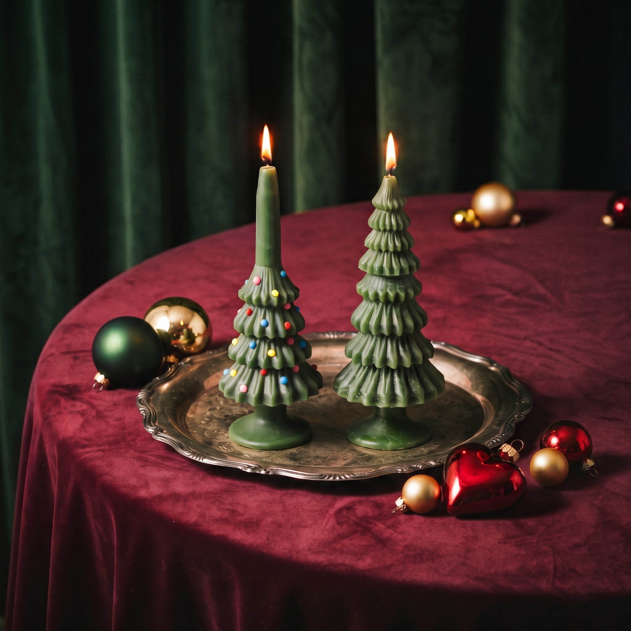 Two Christmas Tree Taper Candles burning on a silver tray with a red velvet tablecloth — one decorated with colorful sprinkle ornaments, one plain green — surrounded by Christmas ornaments.