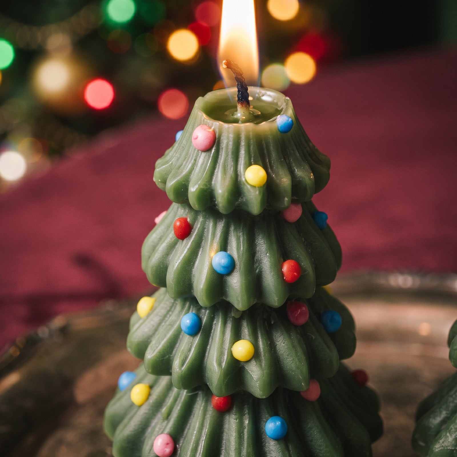Close-up of a lit Christmas Tree Taper Candle with colorful sprinkle-like ornaments pressed into its ridged green layers, glowing string lights blurred in the background.