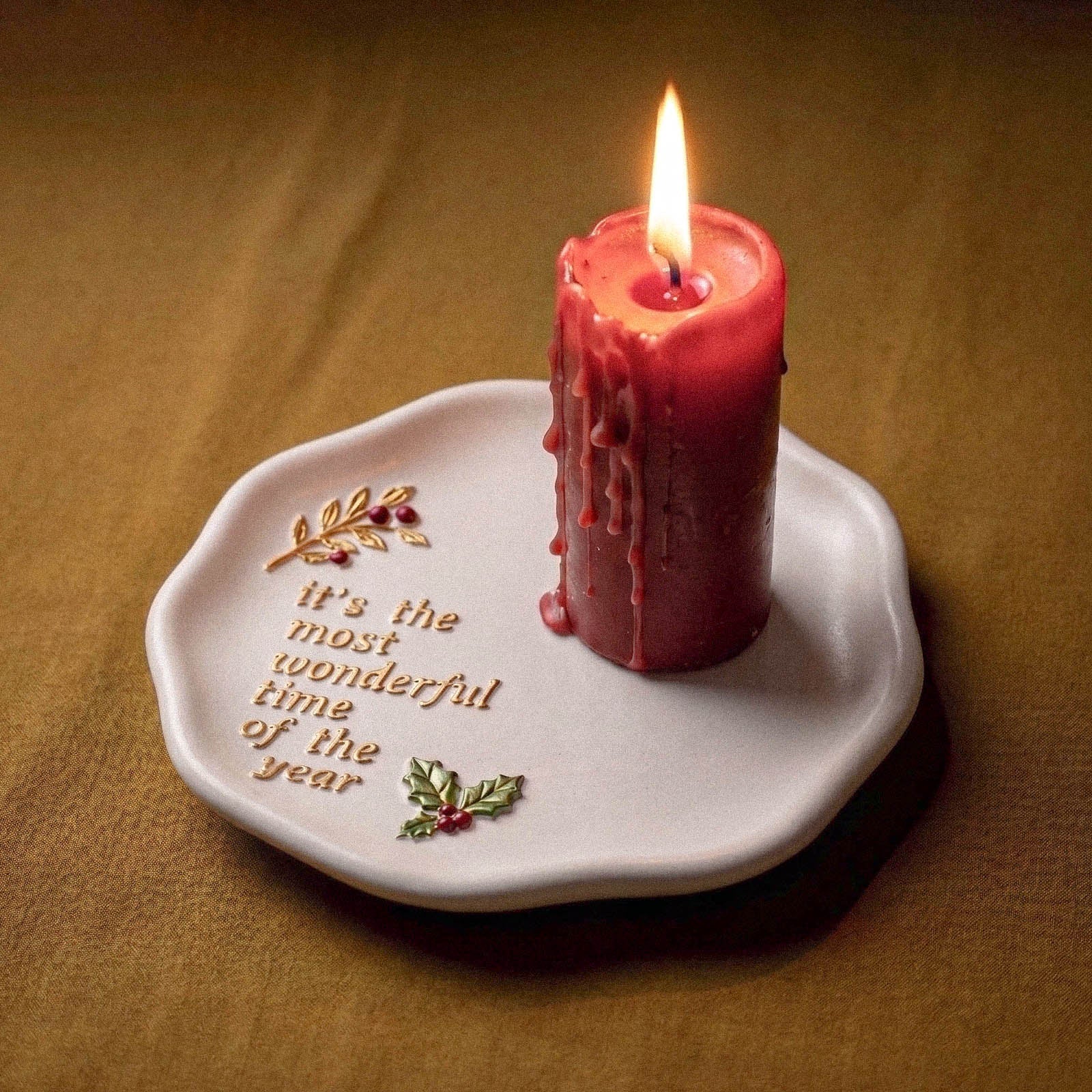 A dripping red pillar candle burning on a white trinket tray embossed with It's the Most Wonderful Time of the Year and a gold holly branch, set against a dark olive backdrop.
