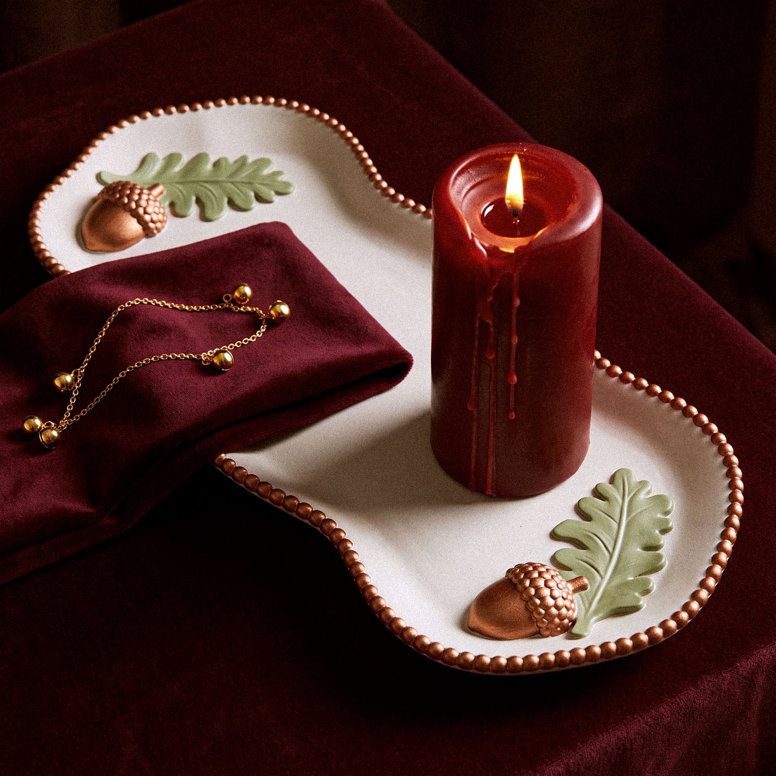 A dripping red pillar candle burning on a Forest Acorn trinket tray beside a folded burgundy velvet napkin and a gold beaded bracelet, styled on dark velvet fabric.