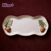 Studio82 x Boowan Nicole Forest Acorn Tray - overhead view of a wavy scalloped white trinket tray with a beaded bronze rim and a sculpted oak leaf and acorn at each end, Nico+ collaboration logo in the corner.