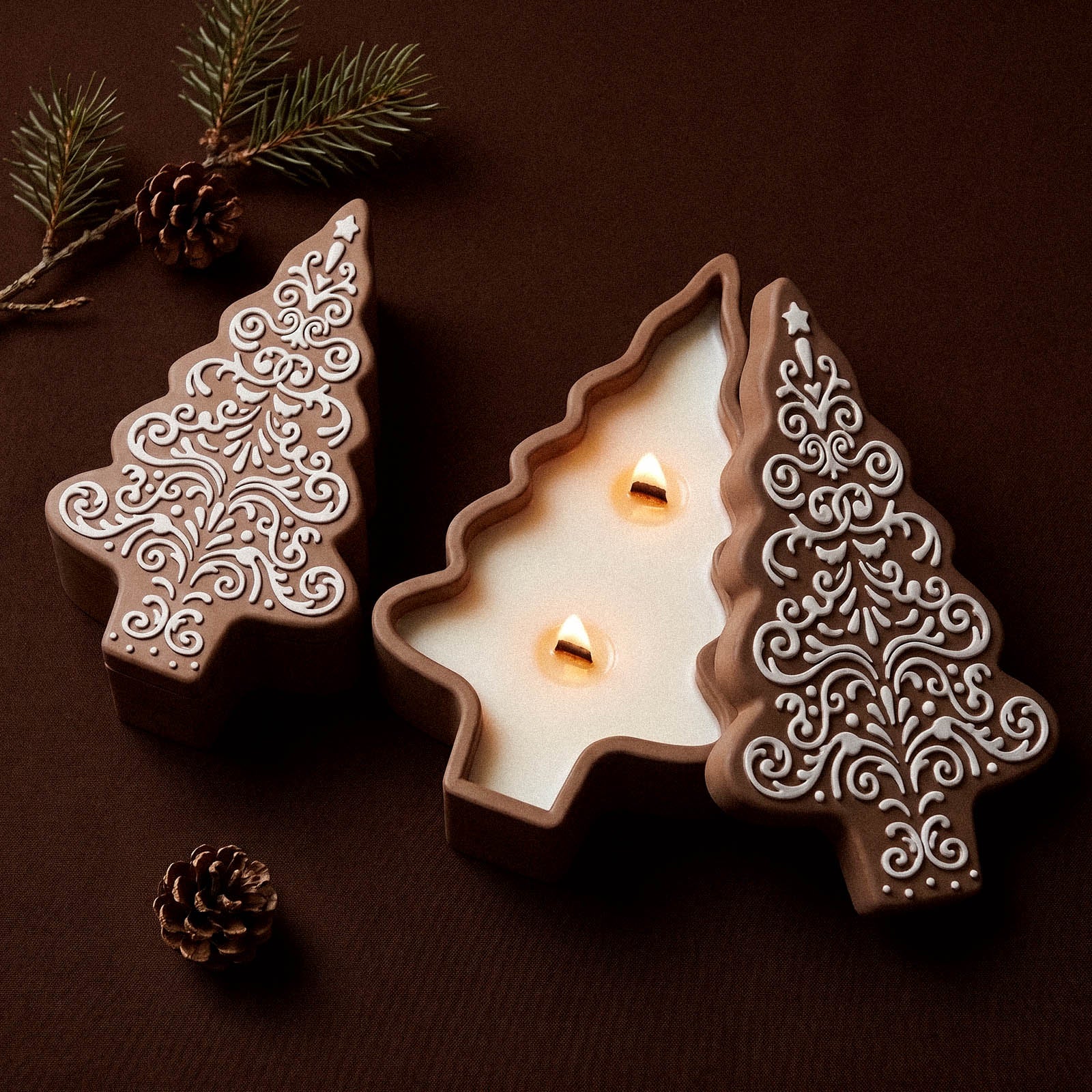 An open gingerbread-lace patterned Christmas tree candle jar with two lit wicks beside a closed matching tree-shaped candle jar, on a dark background with pine sprigs and pinecones.