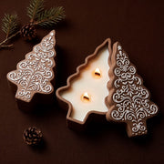 An open gingerbread-lace patterned Christmas tree candle jar with two lit wicks beside a closed matching tree-shaped candle jar, on a dark background with pine sprigs and pinecones.