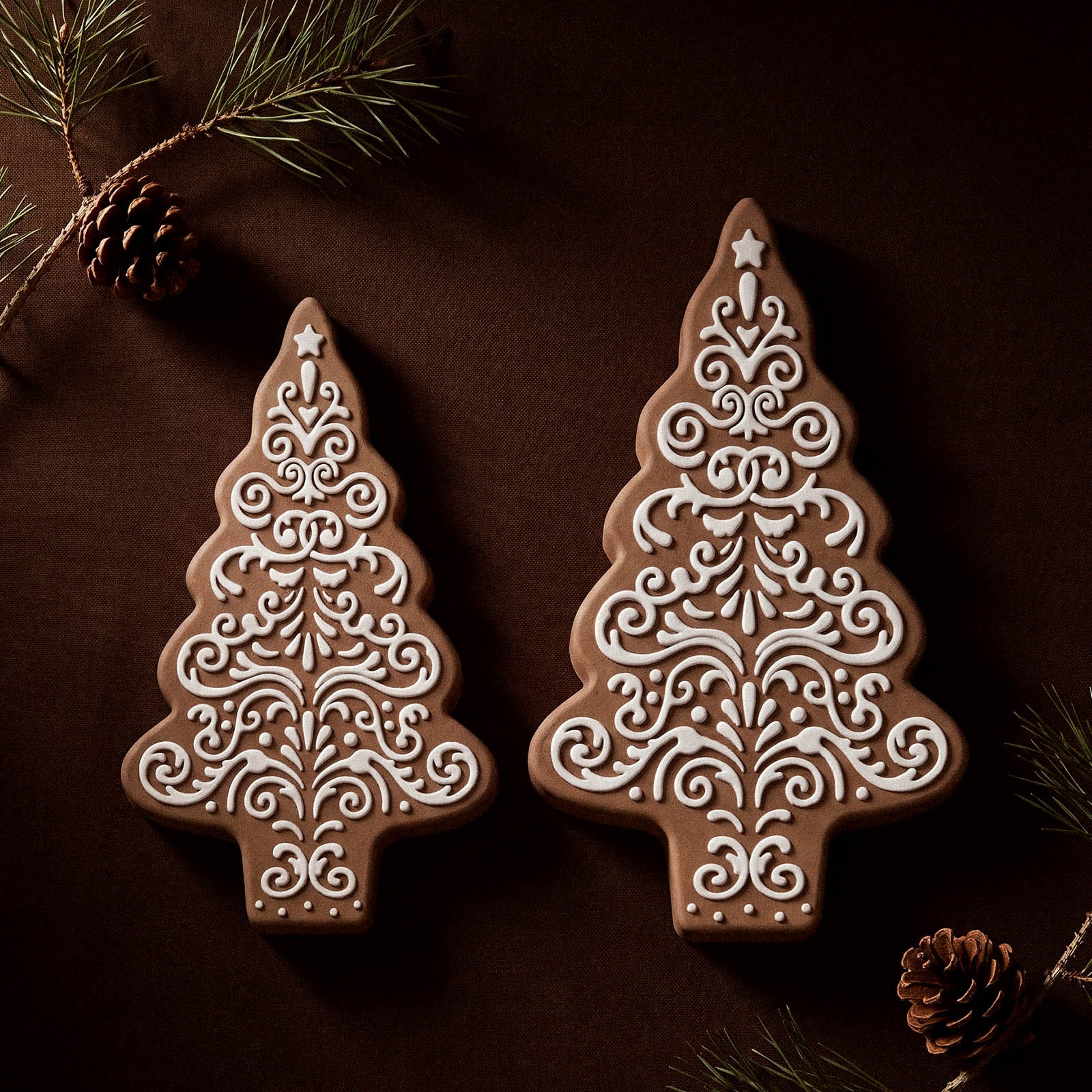 Two gingerbread-lace patterned Christmas tree shaped candle jars in different sizes, displayed side by side on a dark background with pine sprigs and pinecones.