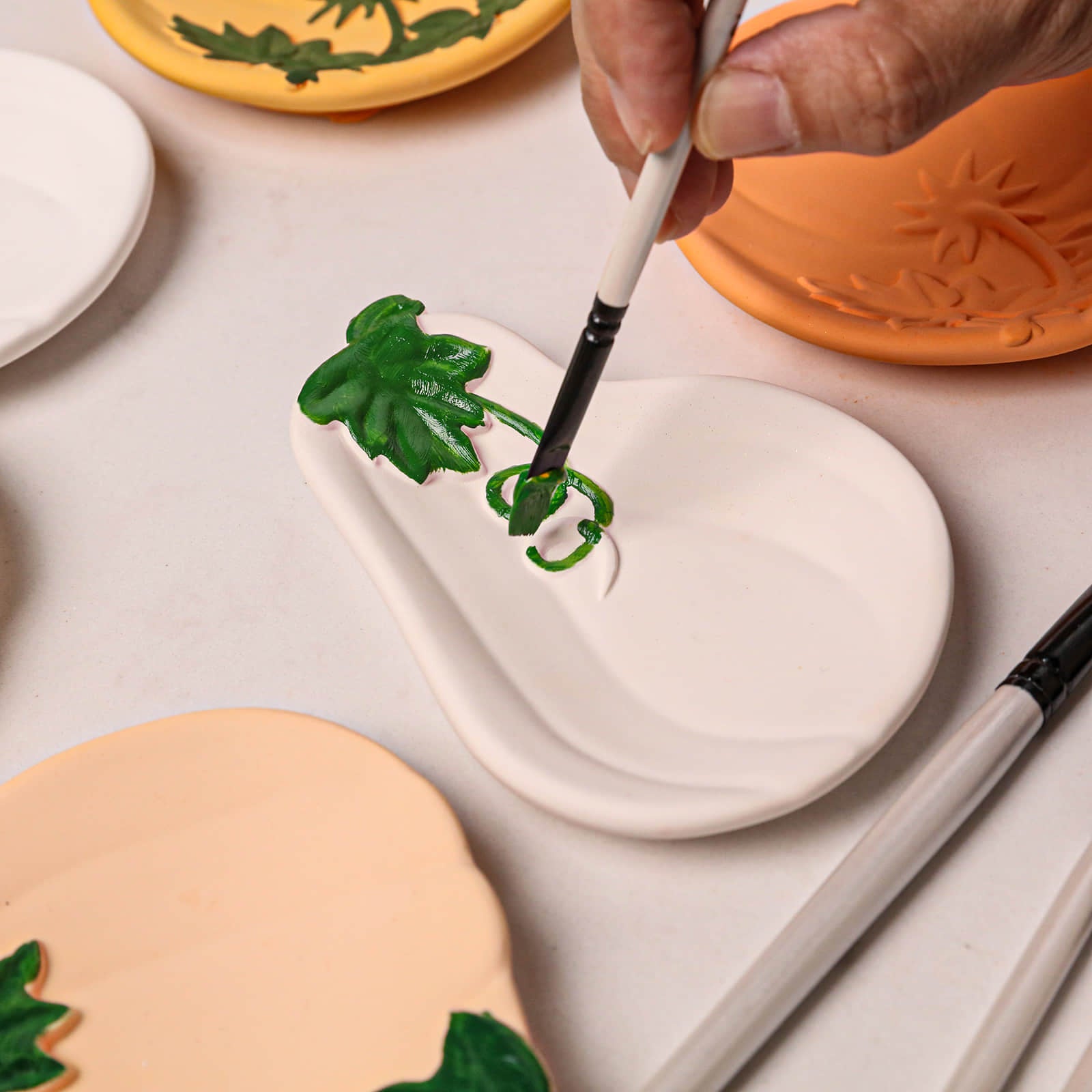 A close-up of a hand painting green leaf details onto a white ceramic plate using a fine paintbrush. Other painted ceramic plates in orange, yellow, and peach are visible in the background on a light surface.