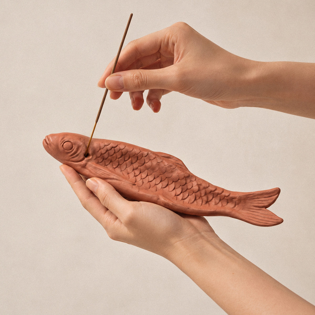 Hands holding a terracotta-coloured fish incense holder while inserting an incense stick