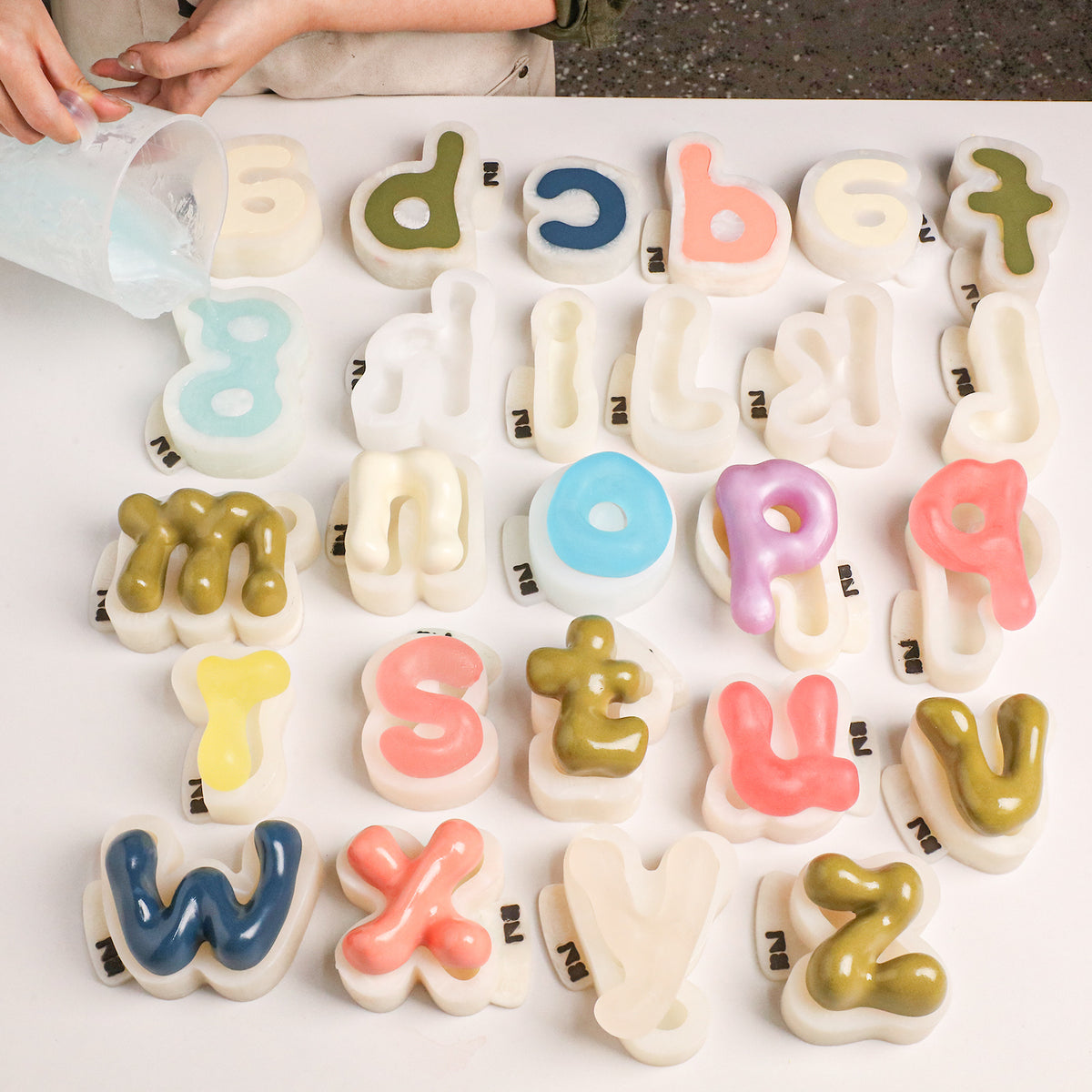 Pouring soap base into lowercase alphabet silicone molds beside finished letter soaps