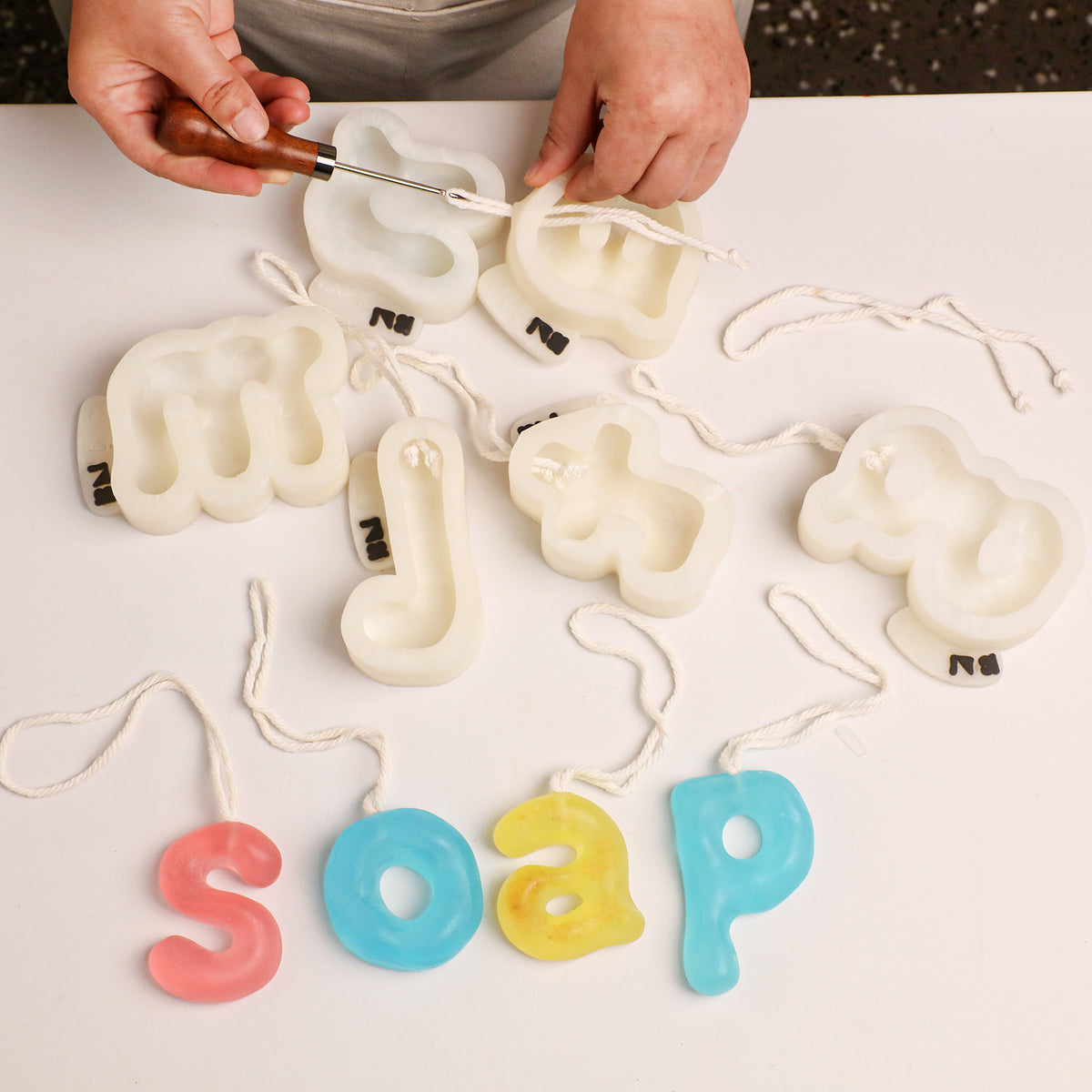 Preparing cord for hanging letter soaps beside silicone molds and letters spelling soap