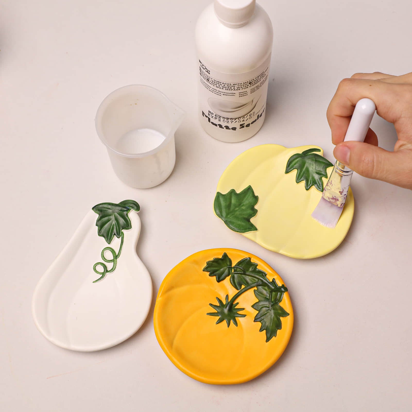 A hand applies white glue from a brush to two decorative ceramic dishes shaped like pumpkins in yellow and orange, alongside a white pear-shaped dish and a small cup of white glue on a light beige surface.