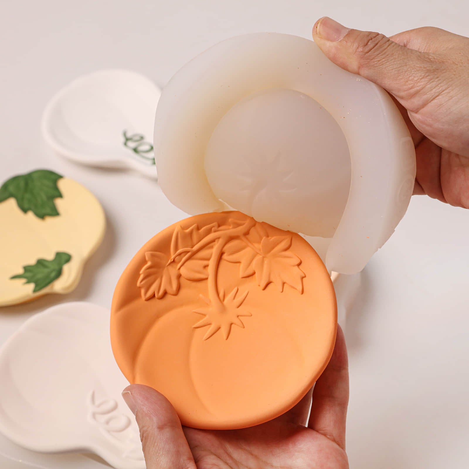 A hand holds an orange silicone mold with leaf and swirl patterns, while another hand lifts a translucent white ring-shaped mold piece above it. A few small decorative soap bars with green leaf designs are visible in the background on a white surface.