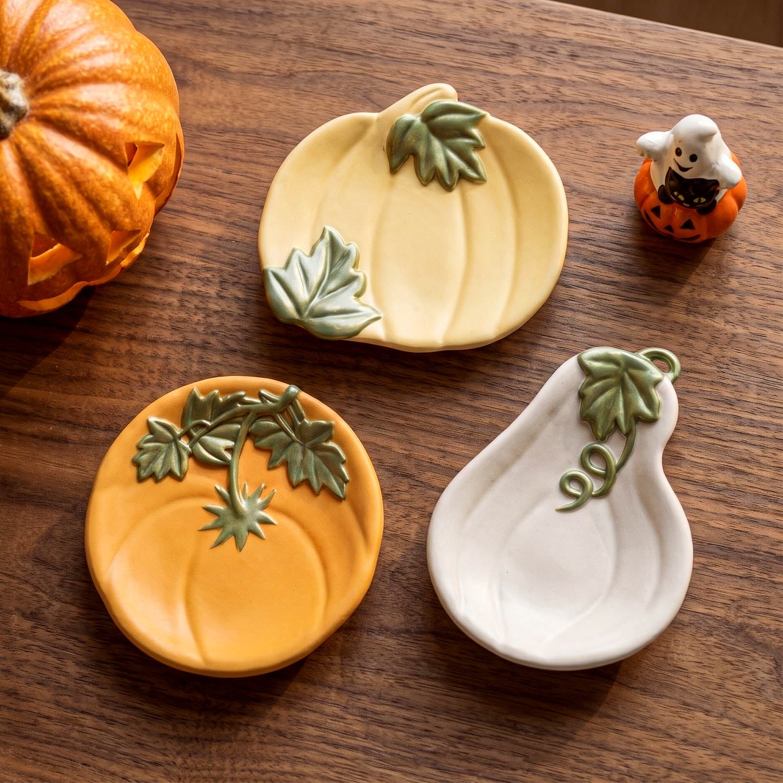 Three decorative ceramic pumpkin-shaped dishes in orange, pale yellow, and white, arranged on a dark wooden table with a carved pumpkin and a small ghost figurine nearby.