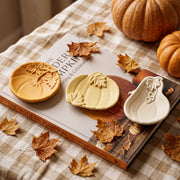 Three hand-painted ceramic pumpkin-shaped dishes in terracotta, cream, and white, arranged on a book titled 'The Modern Pumpkin' with scattered autumn leaves and pumpkins in the background.