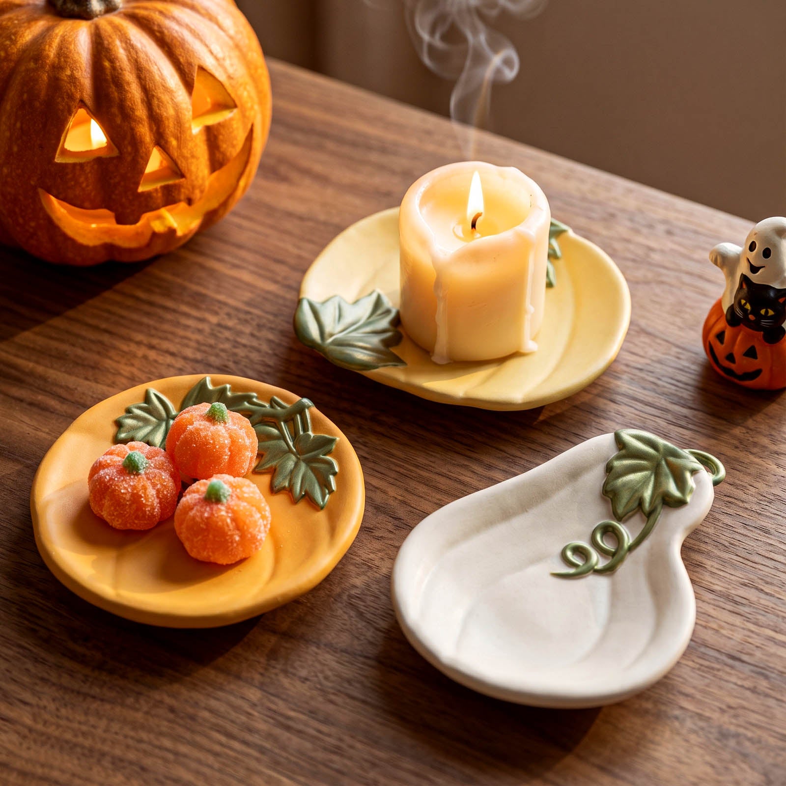 Three ceramic autumn-themed plates on a wooden table: one yellow plate with a green leaf design holding a lit candle, one yellow plate with three small orange pumpkin figures, and one white plate shaped like a pear with a green leaf design. A carved pumpkin and a small ghost decoration are in the background.