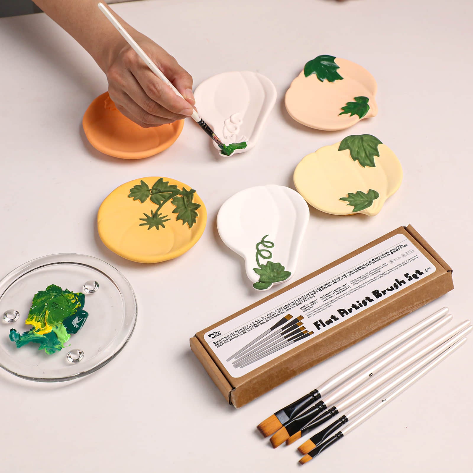 A hand is shown painting on a white ceramic palette with a green paintbrush. Several other painted ceramic palettes in yellow, peach, and cream are arranged on a white surface, along with a brown cardboard box labeled 'Flat Artist Brush Set' and several paintbrushes.