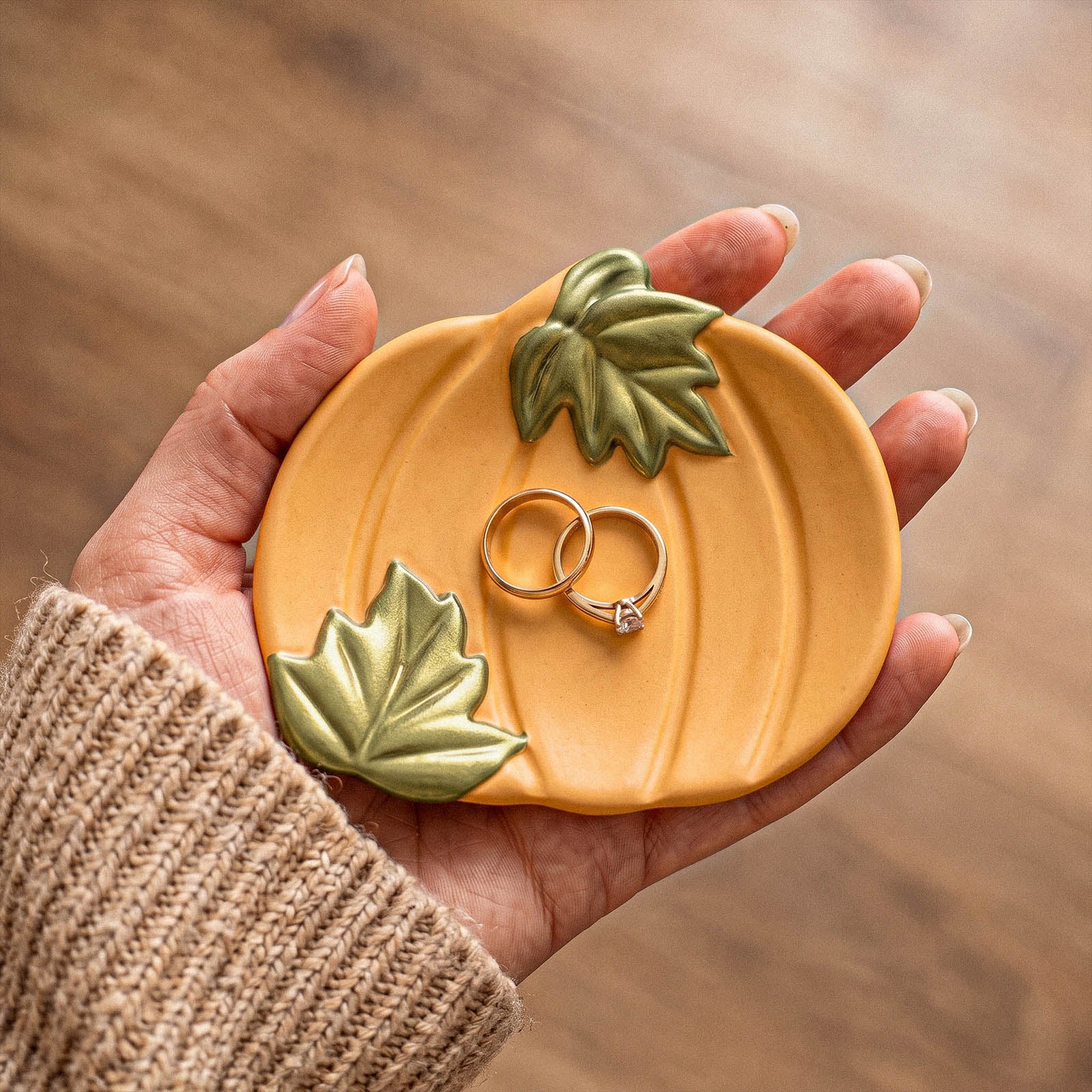 A hand wearing a beige knit sweater holds a pumpkin-shaped ring holder with two gold rings on top. The holder has a warm orange body with green leaf details and is photographed against a blurred wooden background.
