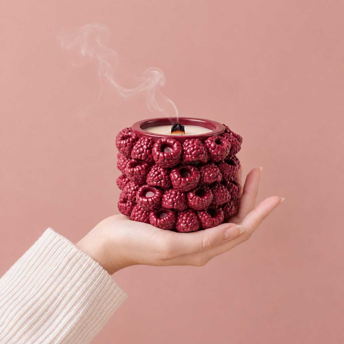 A hand holding a raspberry-textured candle jar against a pink background