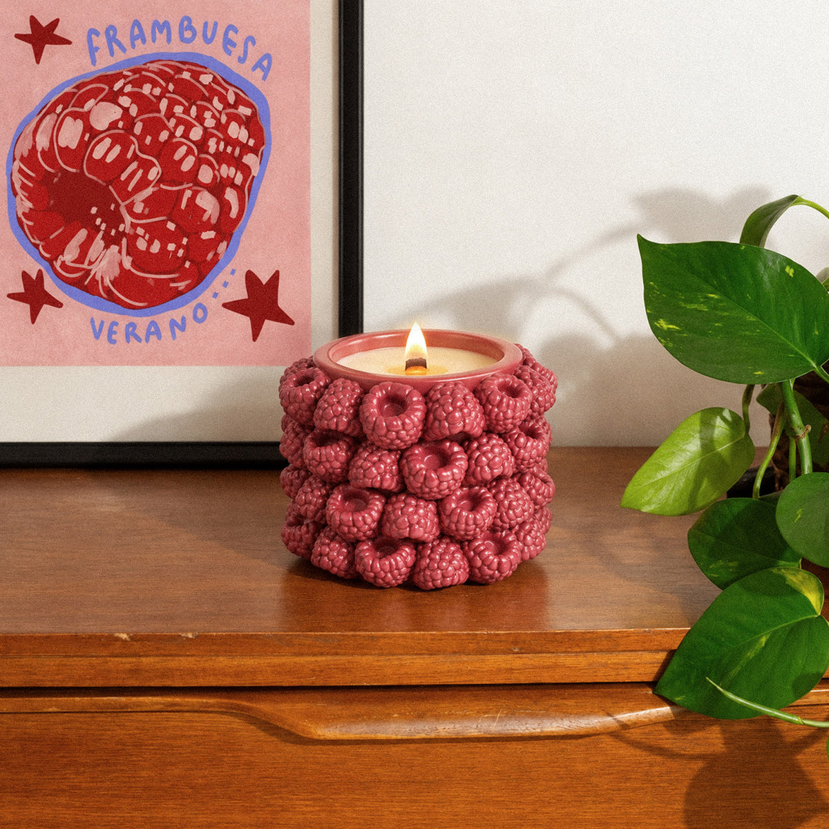 A lit raspberry candle jar on a wooden cabinet beside a plant and framed fruit artwork