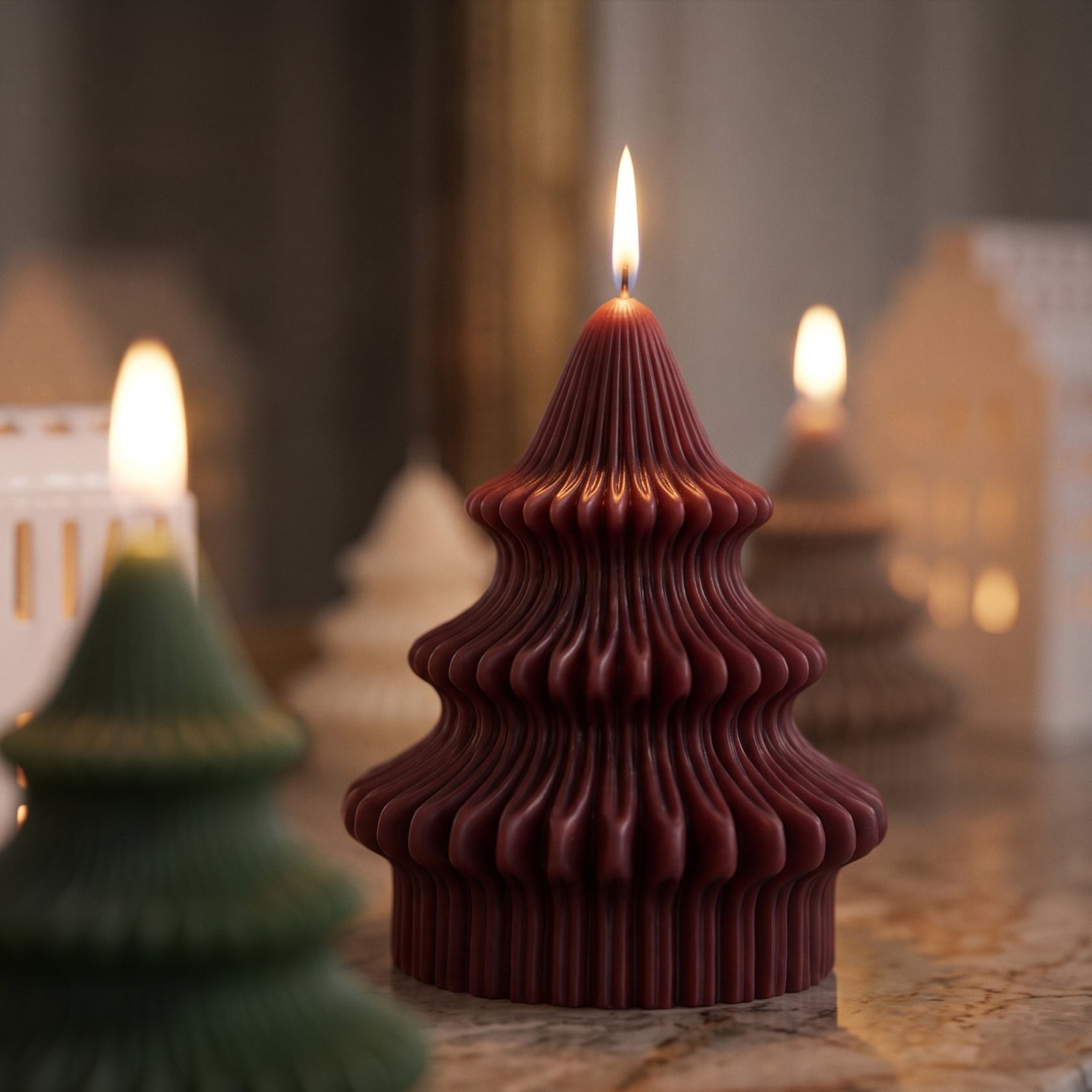 A lit burgundy red Sculpted Christmas Tree Candle with a ribbed three-tier design, displayed among other tree-shaped candles and small house ornaments.