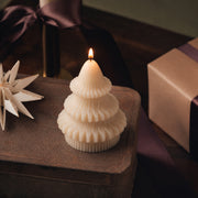 A lit ivory Sculpted Christmas Tree Candle with a ribbed three-tier design on a rustic wooden surface, next to a paper star ornament and wrapped gift boxes with purple ribbon.