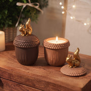 A lit squirrel acorn candle jar on a wooden table styled for Christmas with fairy lights, gold and terracotta squirrel figures nearby.