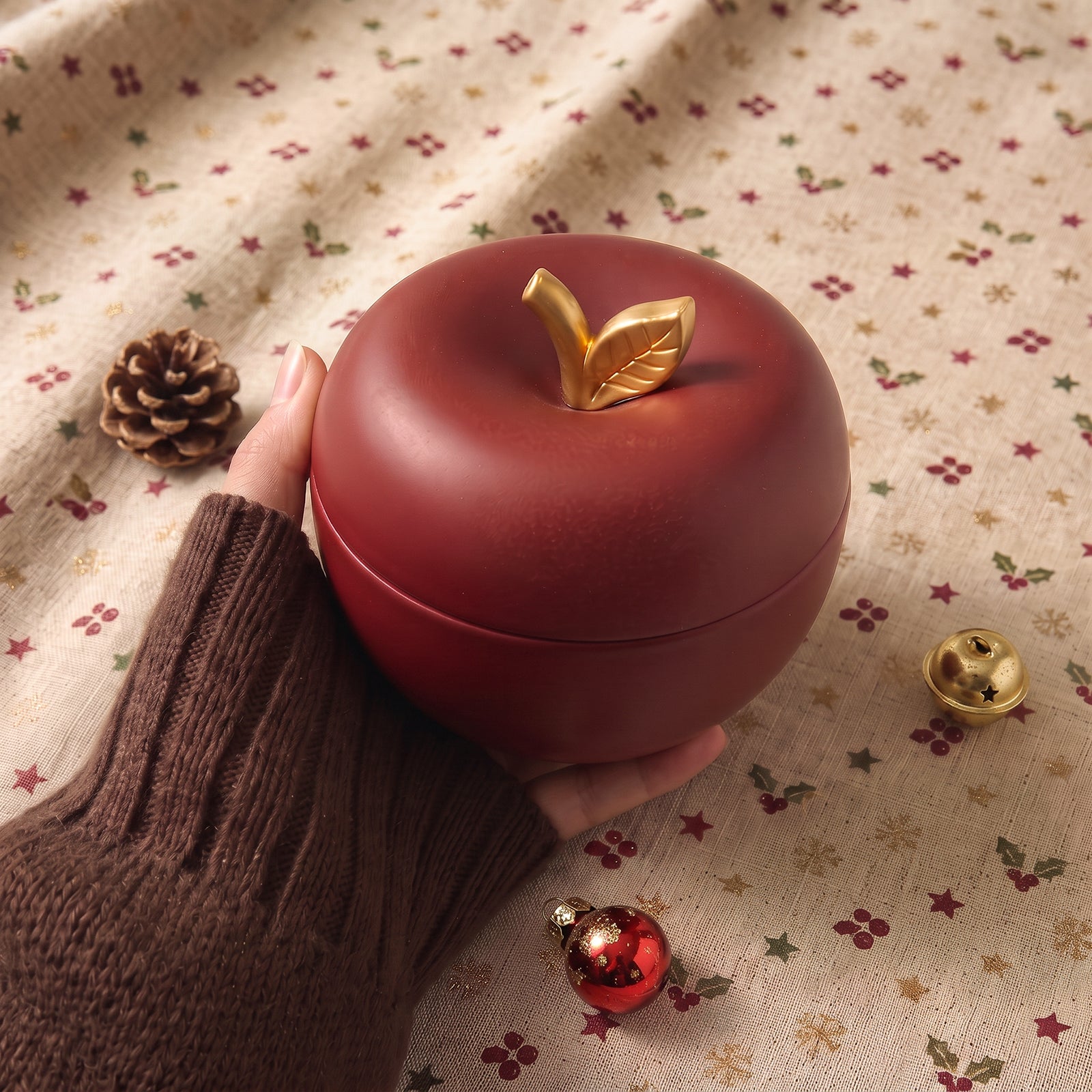 A hand holding a burgundy apple-shaped candle jar with gold leaf lid finial, styled with a pinecone, bell, and Christmas ornament on holiday fabric.