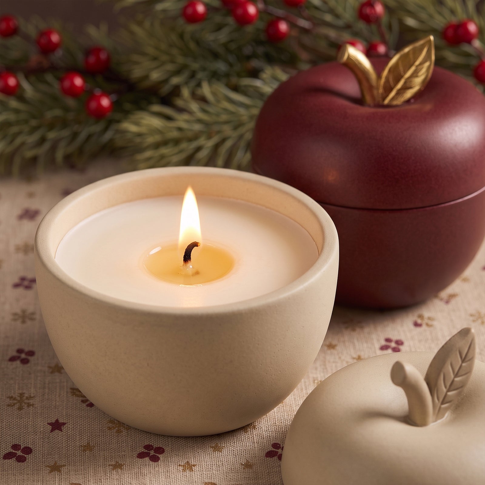 A lit candle burning inside a cream apple-shaped jar next to its burgundy counterpart with gold leaf lid, styled on a festive holiday table linen.
