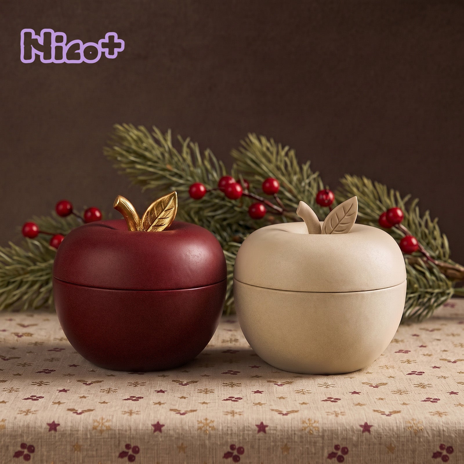 Nico+ lifestyle scene showing two apple-shaped candle jars, one burgundy and one cream, each topped with a gold or matching leaf lid finial, styled with pine and holly berries.