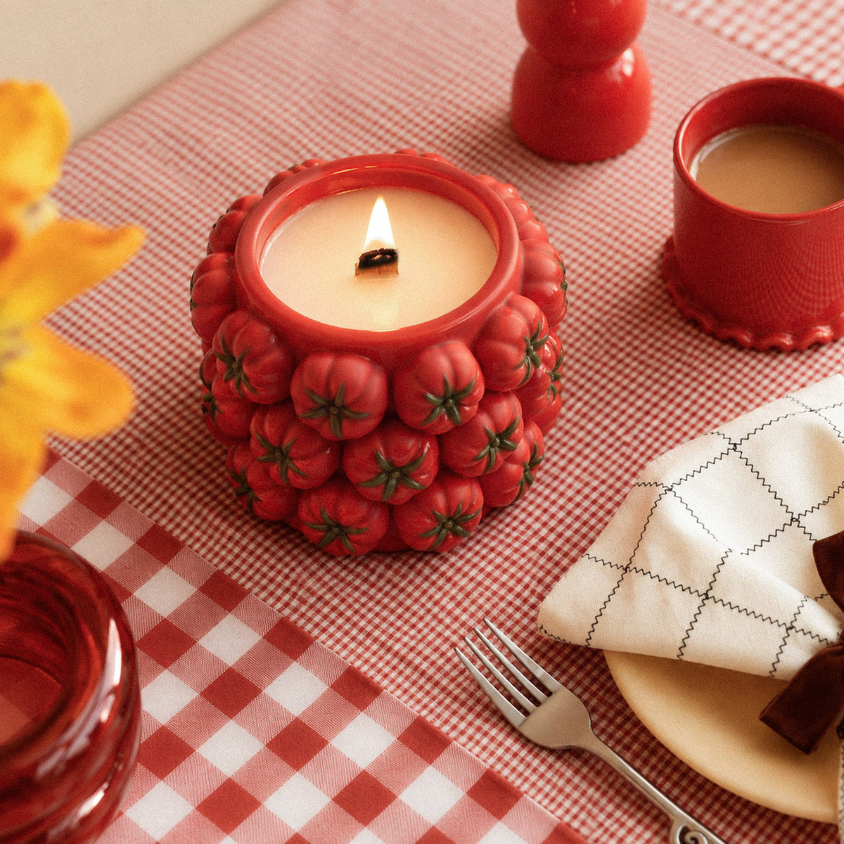 A lit red tomato-cluster candle jar on a red gingham tablecloth