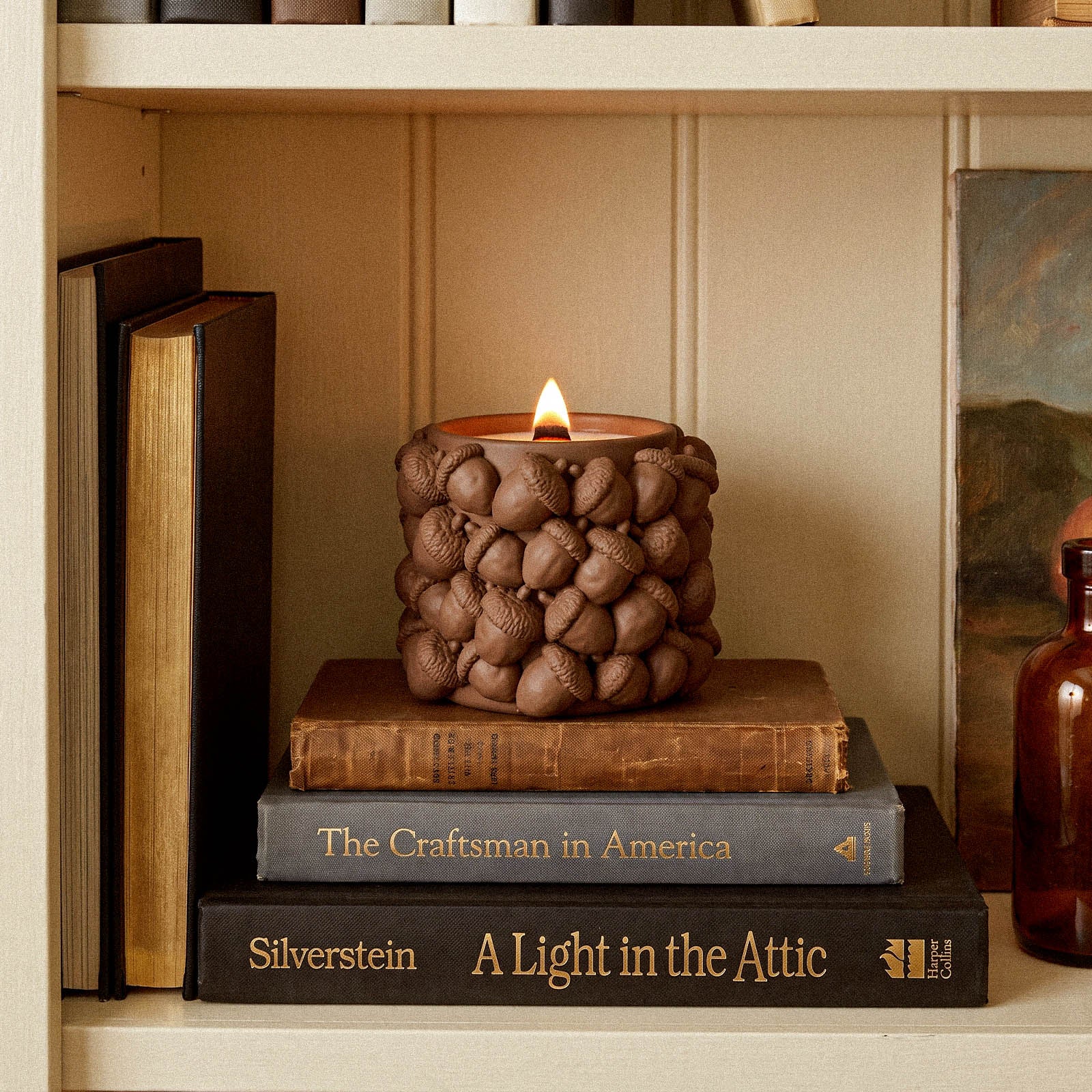 A lit acorn candle jar resting on a stack of books on a bookshelf, styled with a nearby painting and amber glass bottle.