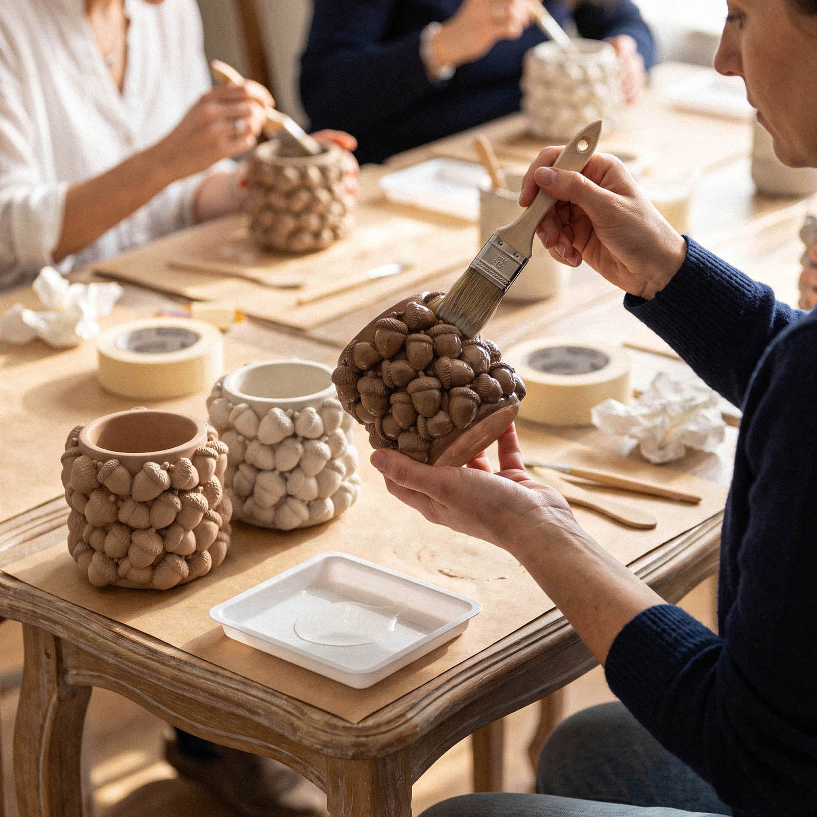 An artisan brushing glaze onto an unfinished acorn candle jar, with raw terracotta and bisque-colored jars on the table.