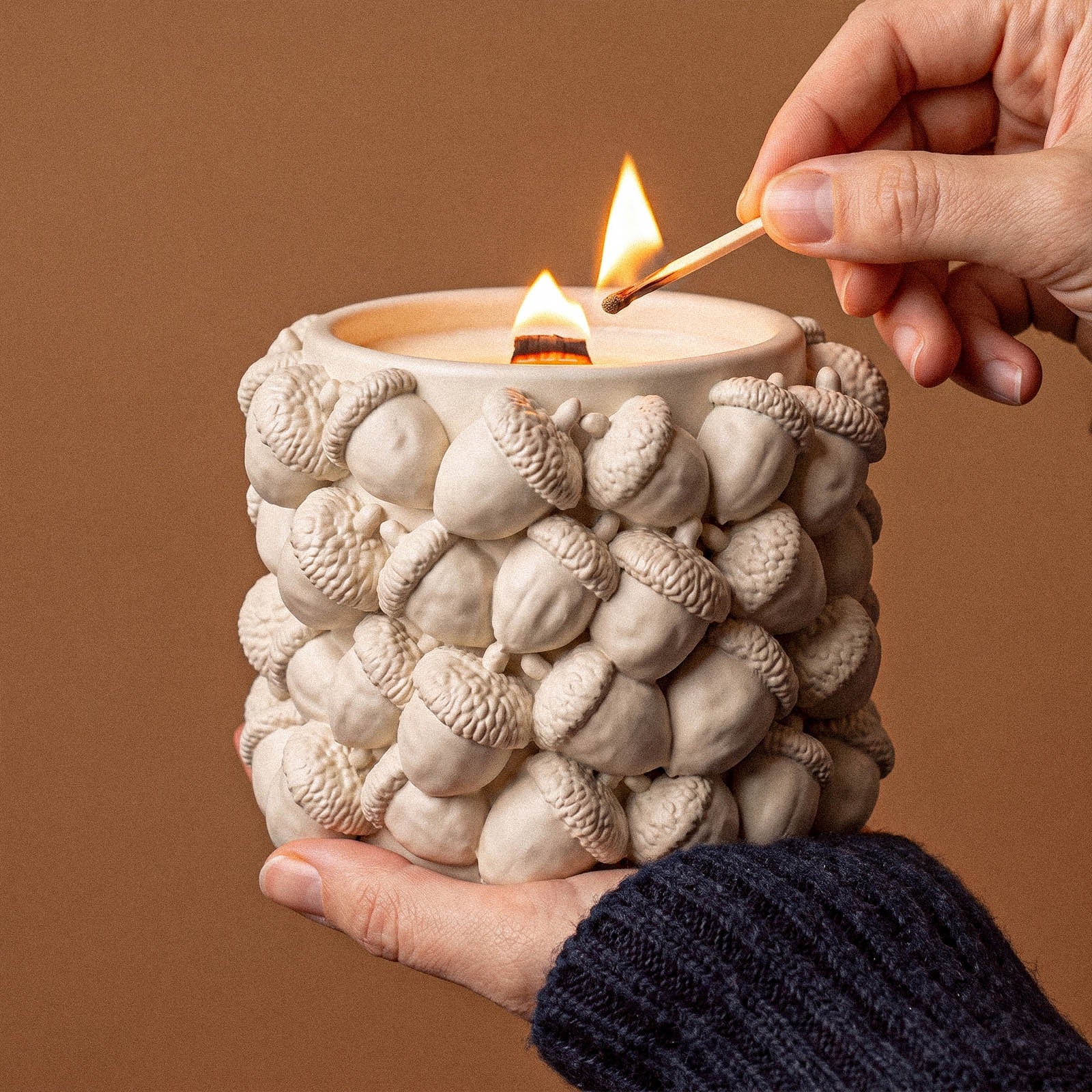 A hand lighting an ivory acorn-textured candle jar with a wooden match against a warm brown background.